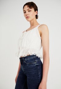 Λευκό, ελαφρύ, αμάνικο τοπ με βολάν από δαντέλα στο τελείωμα και κατακόρυφες ρίγες, συνδυασμένο με σκούρο μπλε τζιν ψηλόμεσο.