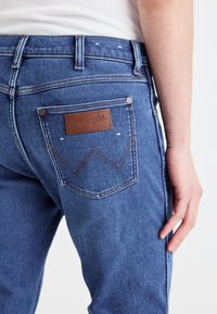 Niebieskie dżinsy z denimu z brązową skórzaną łatą Wrangler, klasyczny design kieszeni, mosiężne nity i niewielkie wyblaknięcia na materiale.