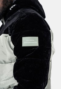 Vadderad jacka med en svart sammetsärm, huvuddel av ljusgrön tyg och en märkespatch med texten "LA ROSA COPENHAGEN."