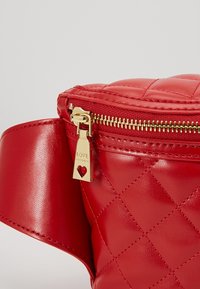 Sac en cuir rouge matelassé avec fermeture éclair dorée et découpe en forme de cœur sur le tire-zip, avec le mot "LOVE" gravé.