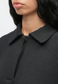 Close-up de uma pessoa a usar uma camisa collared texturada preta com um único botão superior devidamente fechado, mostrando apenas os lábios e o pescoço.
