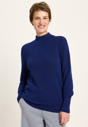 Lächelnde Frau mit kurzen Haaren, die einen blauen Langarmpullover und hellgraue Hosen trägt, mit einer Hand in der Tasche steht.