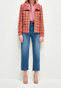 Veste en tartan rose texturé avec col cranté, portée sur un chemisier rose clair, associée à un jean bleu à jambes larges et des talons nude.