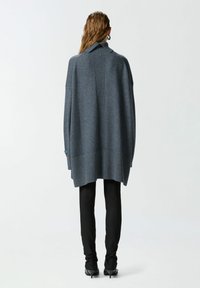 Prevelik tamnosivi pulover s visokim ovratnikom, rebrastom teksturom na rubu i razdvojenim dizajnom na leđima, uparen s crnim hlačama.