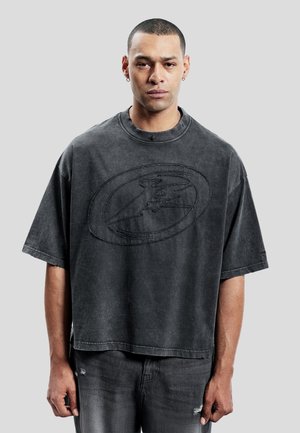 Junger Mann mit kurzen Haaren und Ohrringen, trägt ein weites, ausgebleichtes schwarzes T-Shirt und dunkle, abgenutzte Jeans, steht vor einem schlichten Hintergrund.