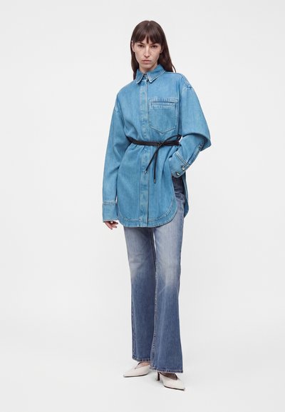 Femme portant une chemise en denim ceinturée surdimensionnée, un jean droit délavé et des talons pointus blancs, debout devant un fond uni.