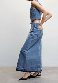 Falda maxi de mezclilla en azul claro con dobladillo desflecado, combinada con un chaleco corto a juego. Tacones negros de punta completan el conjunto.