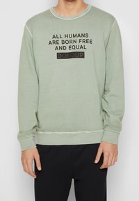 Ljusgrön sweatshirt med ribbat nackband, försedd med svart text som säger "ALLA MÄNNISKOR FÖDS FRIA OCH LIKA" och "DONDUP."