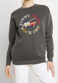 Šedá mikina s kulatým výstřihem a vyšitým vícolor logem: "TOMMY JEANS SINCE 1985" ve zlaté a červené barvě, s žebrovanými manžetami a lemem.