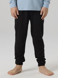 Παιδί που φοράει μαύρο παντελόνι και ανοιχτό μπλε πουκάμισο με μακριά μανίκια, στέκεται ξυπόλυτο σε ουδέτερο γκρι φόντο.