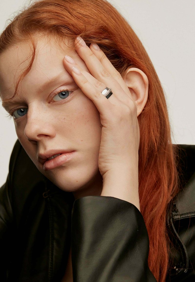 Anillo de metal negro y plateado en una mano apoyada contra una cara, con piel clara y cabello rojo, vistiendo una chaqueta de cuero negra.