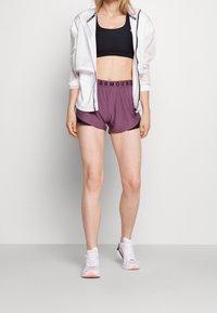 Mujer vistiendo chaqueta blanca con cremallera, sujetador deportivo negro, pantalones cortos morados de Under Armour y zapatillas deportivas blancas, de pie contra un fondo liso.