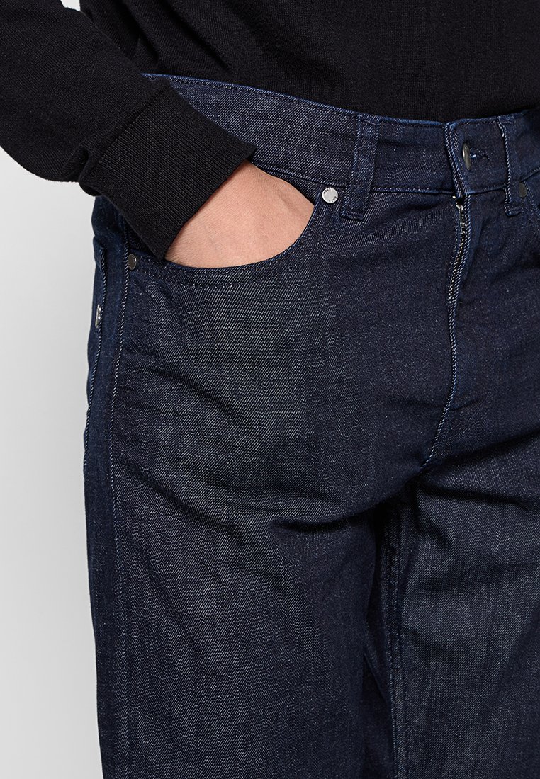 Dunkelblaue Denim-Jeans mit klassischem Design, ausgestattet mit einer Vordertasche, metallischen Hardware-Details und einer strukturierten Stoffoberfläche.