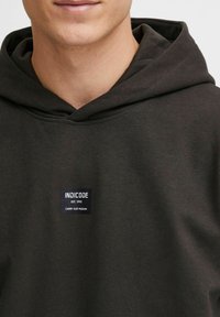 Mørkegrøn hættetrøje lavet af en bomuldsblanding, med en justerbar hætte og et rektangulært mærke med teksten "INDICODE" og "CARRY OUR PASSION".