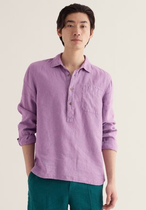 Giovane uomo con capelli scuri che indossa una camicia lavanda a maniche lunghe abbottonata e pantaloni turchesi, in piedi contro uno sfondo chiaro e uniforme.