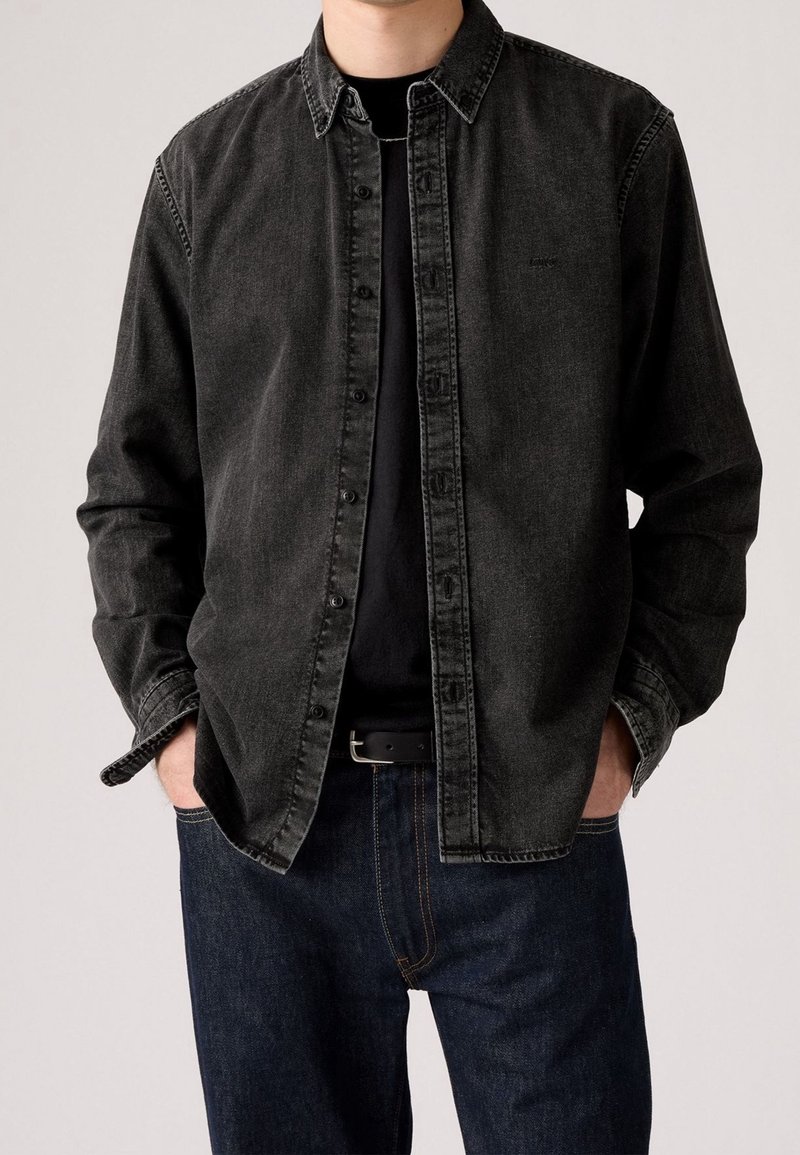 Homme portant une chemise en jean noire par-dessus un t-shirt noir avec un jean bleu foncé et une ceinture noire, mains dans les poches.