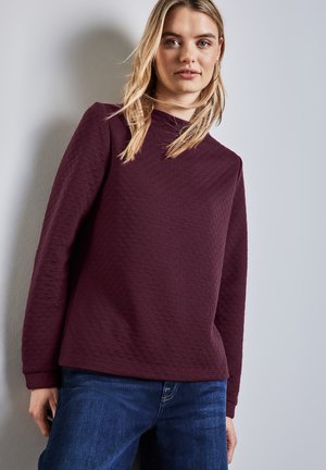 Burgunderfarbener Strickpullover mit strukturiertem Muster, hohem Kragen und langen Ärmeln, kombiniert mit blauen Jeans.