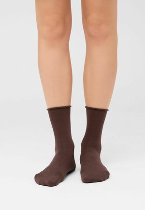 Piernas de una persona que lleva calcetines lisos de color marrón oscuro hasta la mitad de la pantorrilla, de pie sobre una superficie gris claro.
