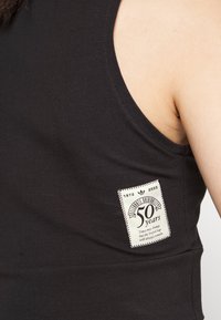 Fekete ujjatlan felső, amelyen nagy, fehér címke található "50 years" felirattal és egy logóval, textúrált anyagból készült.