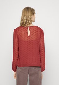 Blusa vermelha transparente com pontos texturizados, mangas longas e detalhe de abertura nas costas. Apresenta um decote redondo e um corte ligeiramente solto.