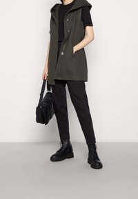 Persona con chaleco sin mangas de color verde oscuro con capucha sobre camiseta negra, pantalones negros, botines negros con cordones y sosteniendo un bolso negro acolchado.
