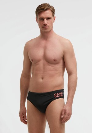 Shirtless man wearing black swim briefs with red "CAMP DAVID" logo on left hip, standing against a plain light background.