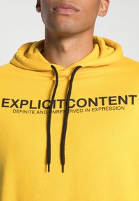 Żółta bluza z kapturem i sznurkiem, z czarnym nadrukiem: "EKSPLICYTNE TREŚCI DEFINITYWNE I BEZ ZASTRZEŻEŃ W WYRAŻENIU." Miękka faktura materiału.
