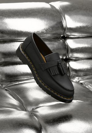 Mocasin de piel negra con borlas y costuras amarillas sobre una superficie metálica plateada brillante y acolchonada.