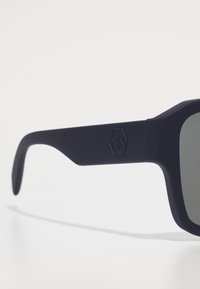 Occhiali da sole blu navy con montatura in plastica opaca, lenti rettangolari e logo inciso discretamente sulla tempia. Texture liscia in tutto.