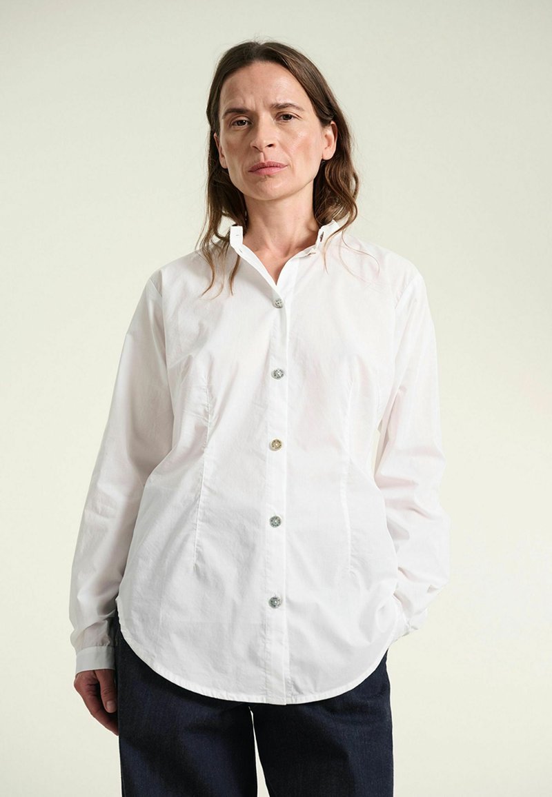 Femme aux longs cheveux bruns portant une chemise blanche boutonnée et un pantalon foncé, debout avec une main dans la poche sur fond clair.