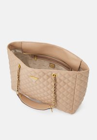 Sac fourre-tout matelassé beige avec des accents de chaîne dorée et une poignée plate. L'intérieur comprend une poche zippée et est doublé d'un motif logo.