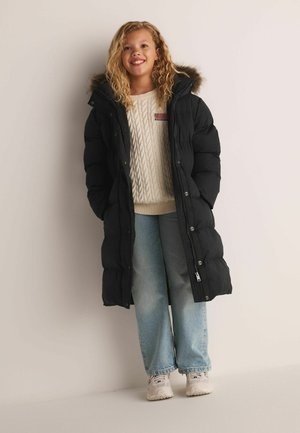 Smiling girl with curly blonde hair wears long black puffer coat with fur hood, cream cable knit sweater, light blue jeans, and white sneakers.
