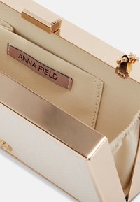Pochette crème ouverte avec cadre métallique doré et étiquette « Anna Field » cousue à l'intérieur sur une doublure en tissu beige.