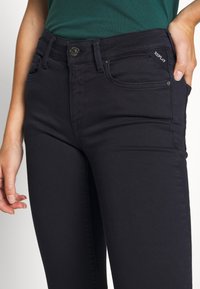 Jeans de mezclilla oscura con corte ajustado, que cuentan con un cierre de botón de metal, bolsillos y una pequeña etiqueta de la marca en la parte frontal.
