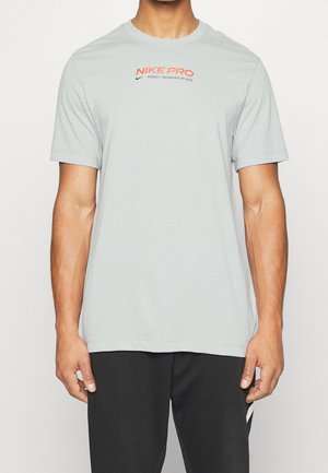 Homme portant un t-shirt Nike Pro gris clair à manches courtes avec un logo orange et un pantalon noir, debout devant un fond uni.