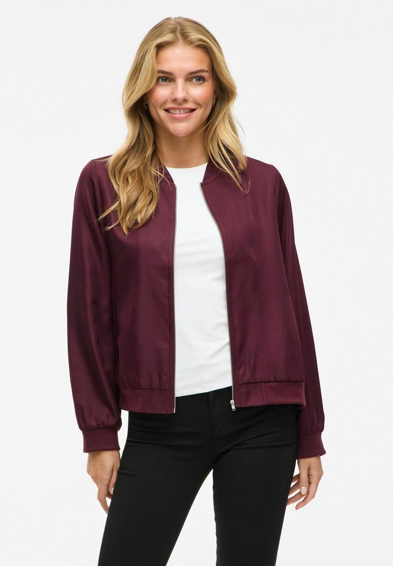 Burgunderfarget sateng bomberjakke med glidelås foran, ribbestrikkede mansjetter og nederkant; brukt over en hvit topp og svarte bukser. Glatt tekstur, avslappet passform.