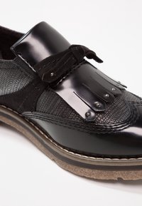 Chaussure habillée en cuir noir avec des panneaux texturés, des franges décoratives et des clous, un détail en suède en forme de nœud, et une semelle en caoutchouc beige sur fond blanc.