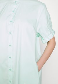 Vêtement à manches courtes et à revers de couleur vert menthe clair, avec des poches latérales, fabriqué en tissu lisse. Présente une coupe décontractée et un subtil éclat.