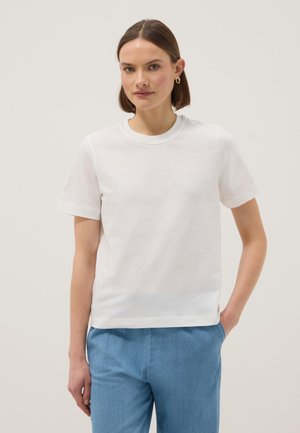 Jeune femme aux cheveux bruns courts portant des boucles d'oreilles créoles dorées, un t-shirt blanc à manches courtes et un pantalon bleu clair, debout avec une main dans la poche.