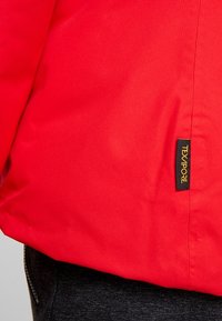 Section de tissu rouge d'une veste avec une étiquette TEXAPORE noire et jaune près d'une couture verticale, associée à un jean en denim foncé visible.