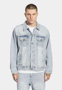 Jasnoniebieska dżinsowa kurtka o luźnym kroju, z dwoma kieszeniami na piersi, zapięciem na guziki i minimalnymi przetarciami.