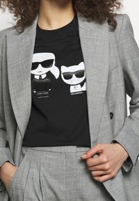 Šedý blazer přes černé tričko s bílými kreslenými postavičkami. Design obsahuje sluneční brýle a stylizované prvky. Text zní "KARL LAGERFELD."