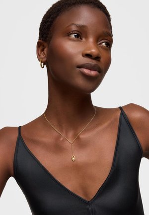 Kobieta o krótkich włosach, nosząca małe złote kolczyki w kształcie kółek, delikatny złoty naszyjnik i czarny bezrękawnik z dekoltem w serek, patrząca nieco na bok.