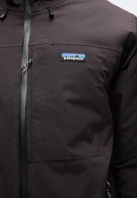 Κοντινό πλάνο μαύρου μπουφάν Patagonia που δείχνει τσέπη στο στήθος με λογότυπο και φερμουάρ μπροστά με μαύρο και λευκό κορδόνι.
