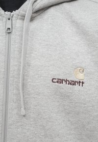 Siva zip-up dukserica od mekog materijala. Sadrži Carhartt logo u tamnocrvenoj i bežoj vezanoj boji. Kapuljača s uzicom i zatvaračem.