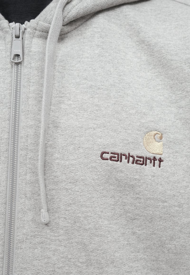Grau gesteppter Kapuzenpullover aus weichem Stoff. Mit einem Carhartt-Logo in dunkelrot und beige gestickt. Kapuze mit Kordelzug und Reißverschluss.