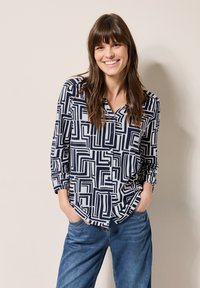 Blusa de patrón geométrico en azul marino y blanco con escote en V, mangas largas y botón frontal, combinada con pantalones de mezclilla azul de corte relajado.