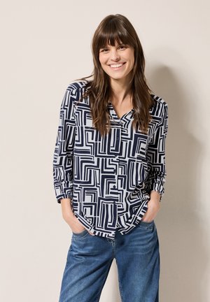 Marineblauwe en witte geometrisch patroon blouse met een V-hals, lange mouwen en kn open voorkant, gecombineerd met relaxte blauwe spijkerbroek.