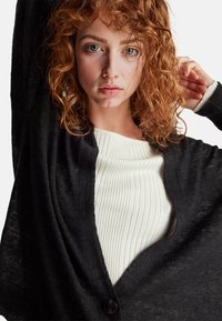 Černý kardigan volného střihu s dlouhými rukávy, zapínáním na knoflíky a lehkou texturou, který je navrstvený na bílém žebrovaném topu.