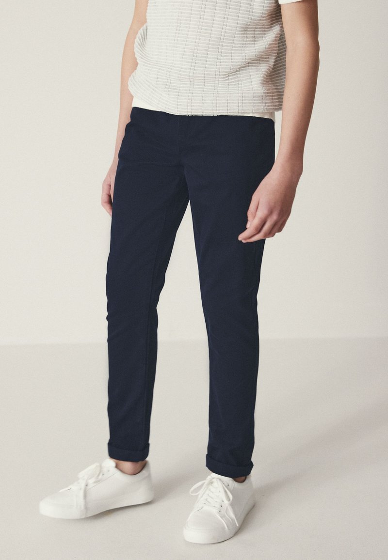 Personne portant un pantalon slim bleu marine foncé, des baskets basses blanches et une chemise blanche texturée à manches courtes sur un fond uni.
